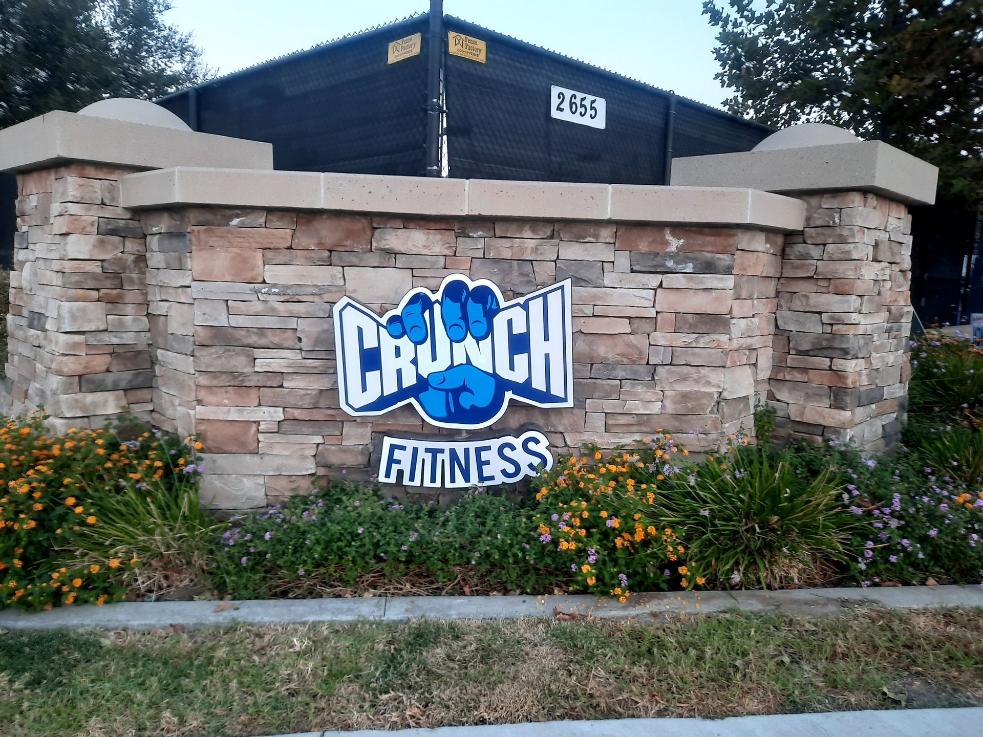 Crunch Fitness sign on a stone wall with blue lettering, in front of a black building and landscaping.