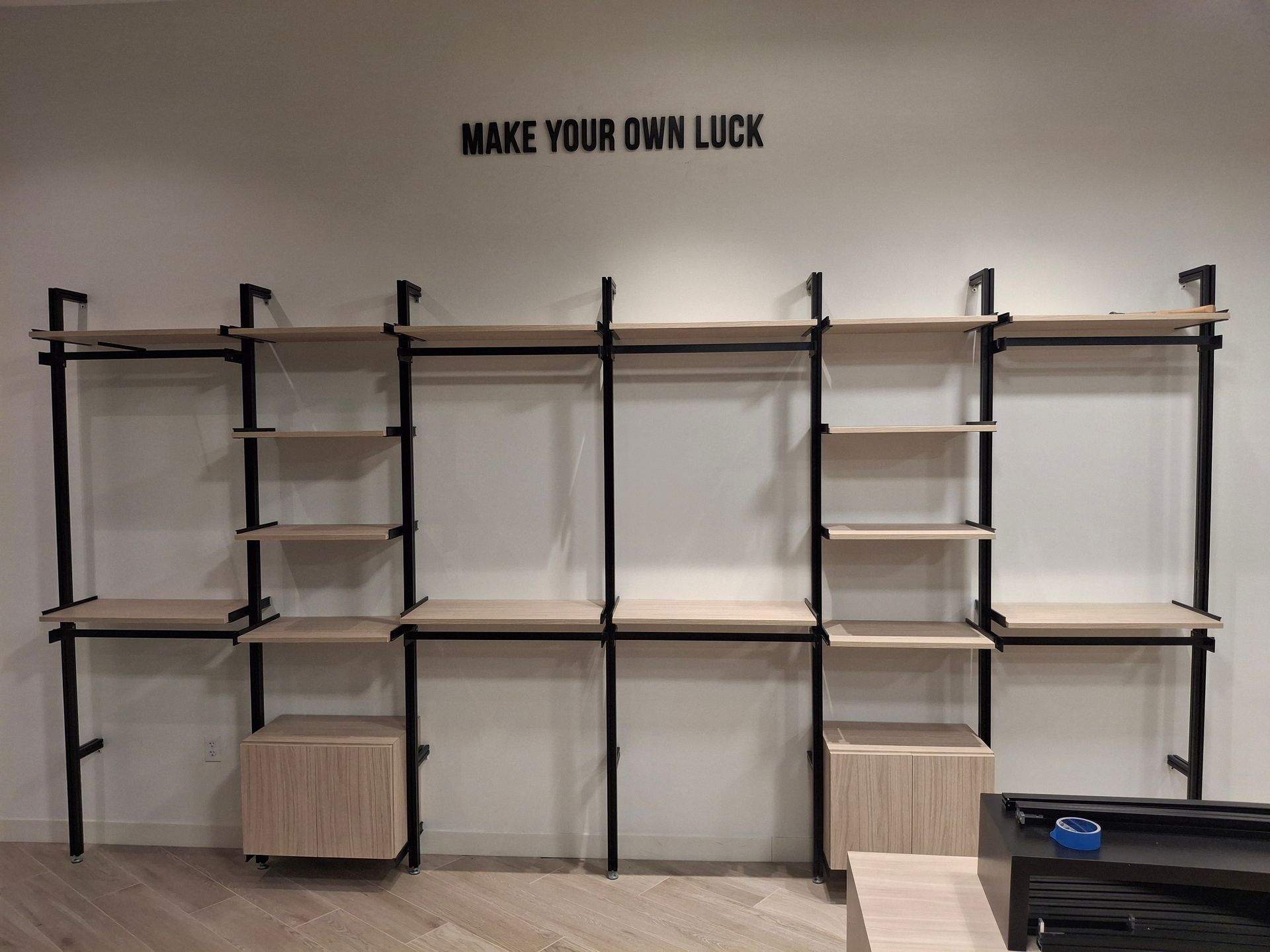 Empty wall-mounted shelving units with black metal frames and wood shelves under the words “MAKE YOUR OWN LUCK.”