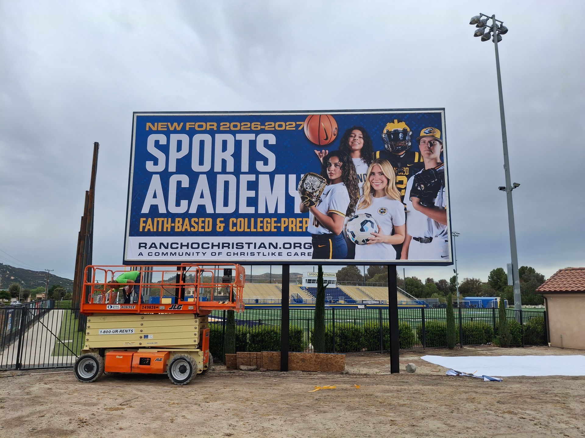 Billboard for sports academy with athlete photos, near an orange lift and construction site under cloudy sky