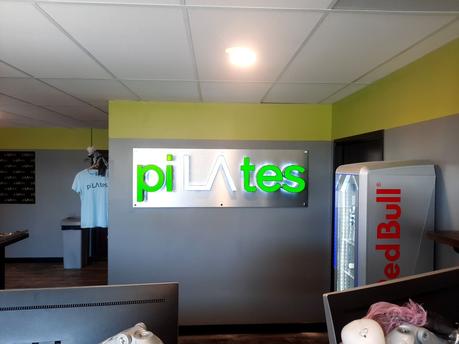 Pilates studio reception with glowing green “pilates” sign and Red Bull fridge