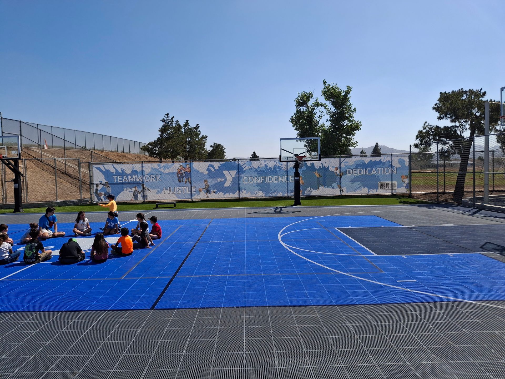 Children seated on a blue sports court outdoors near a chain-link fence and mural