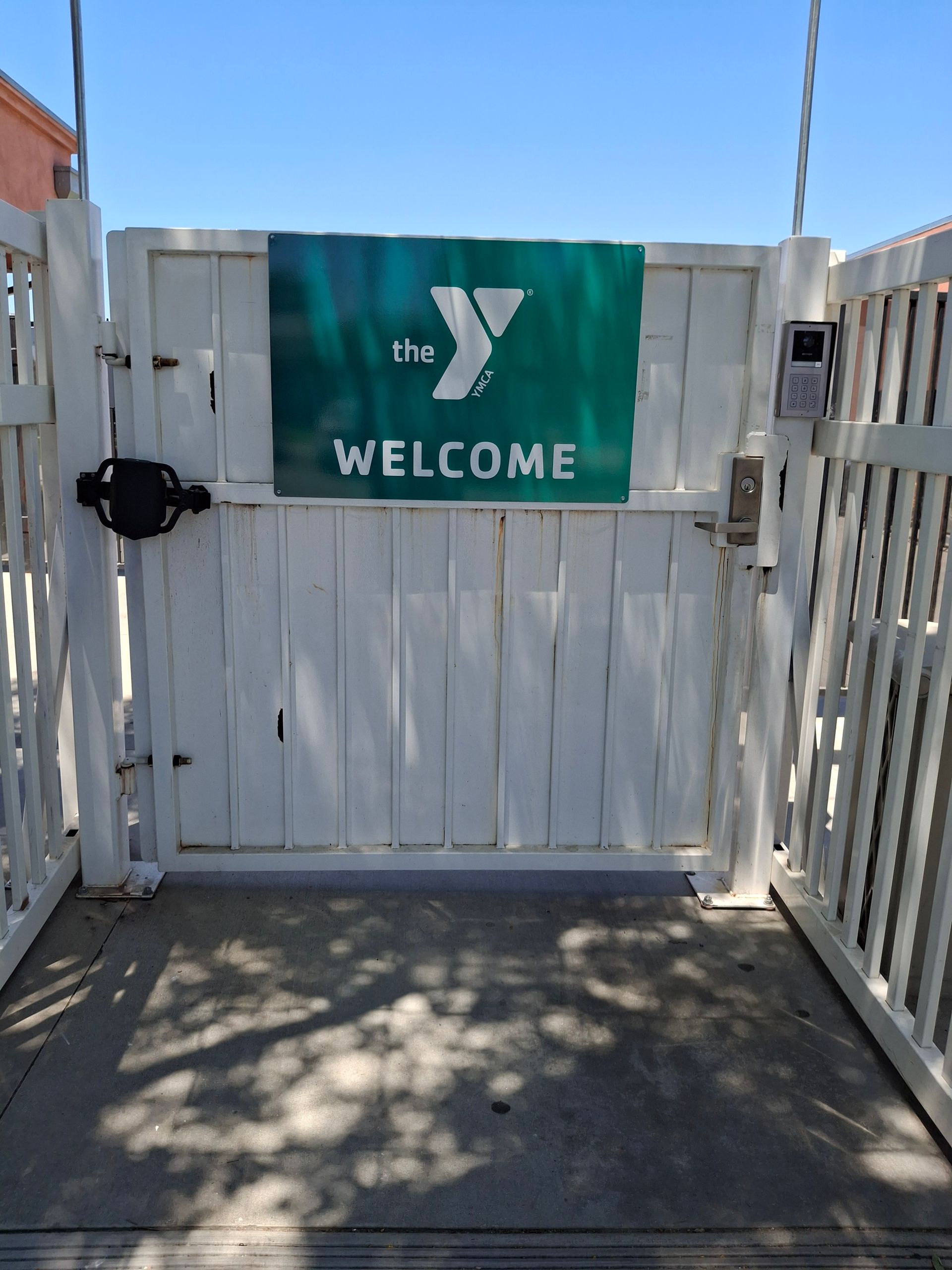 White wooden gate with a green YMCA welcome sign in a sunny outdoor entrance area