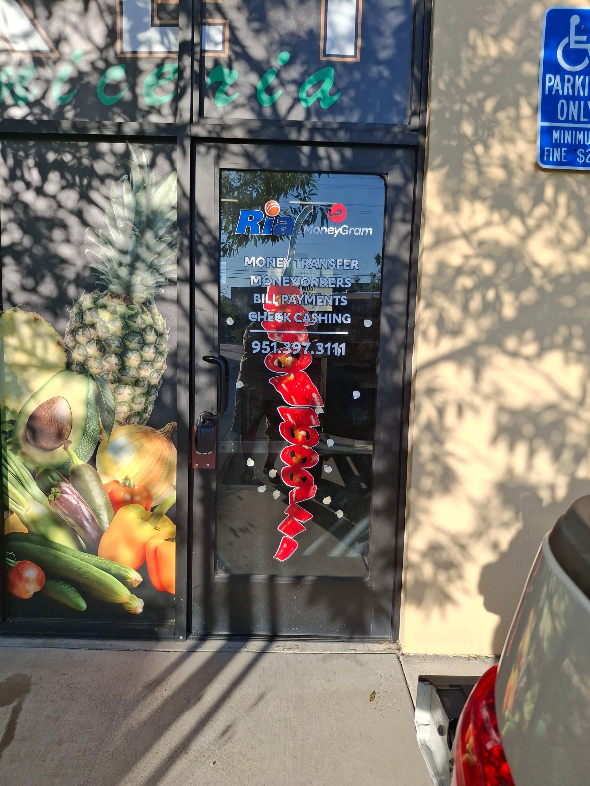 Storefront door with colorful fruit display, red peppers hanging, and a blue accessible parking sign nearby