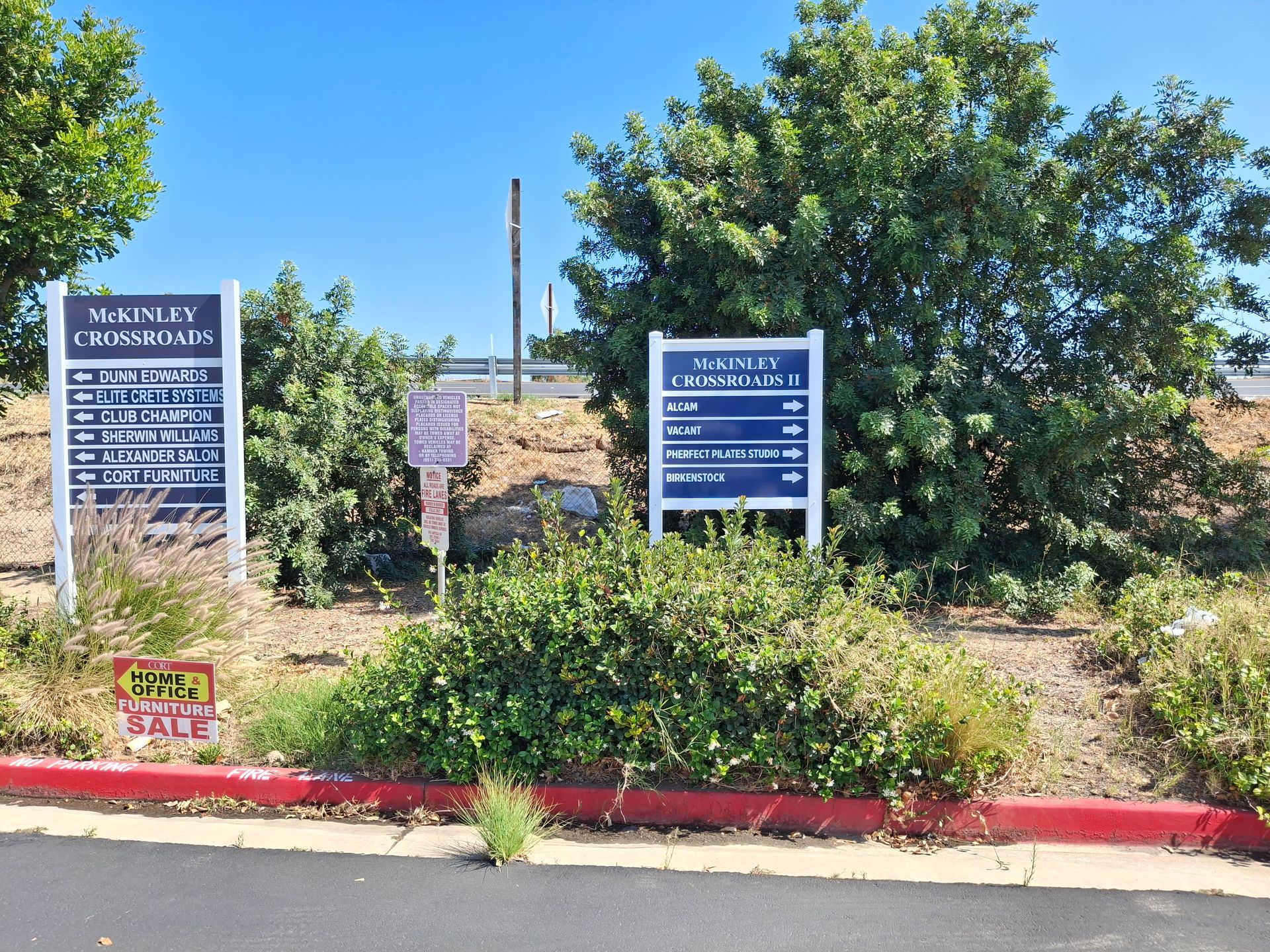 Entrance signs beside a landscaped sidewalk, with blue directional signs and trees under a clear sky