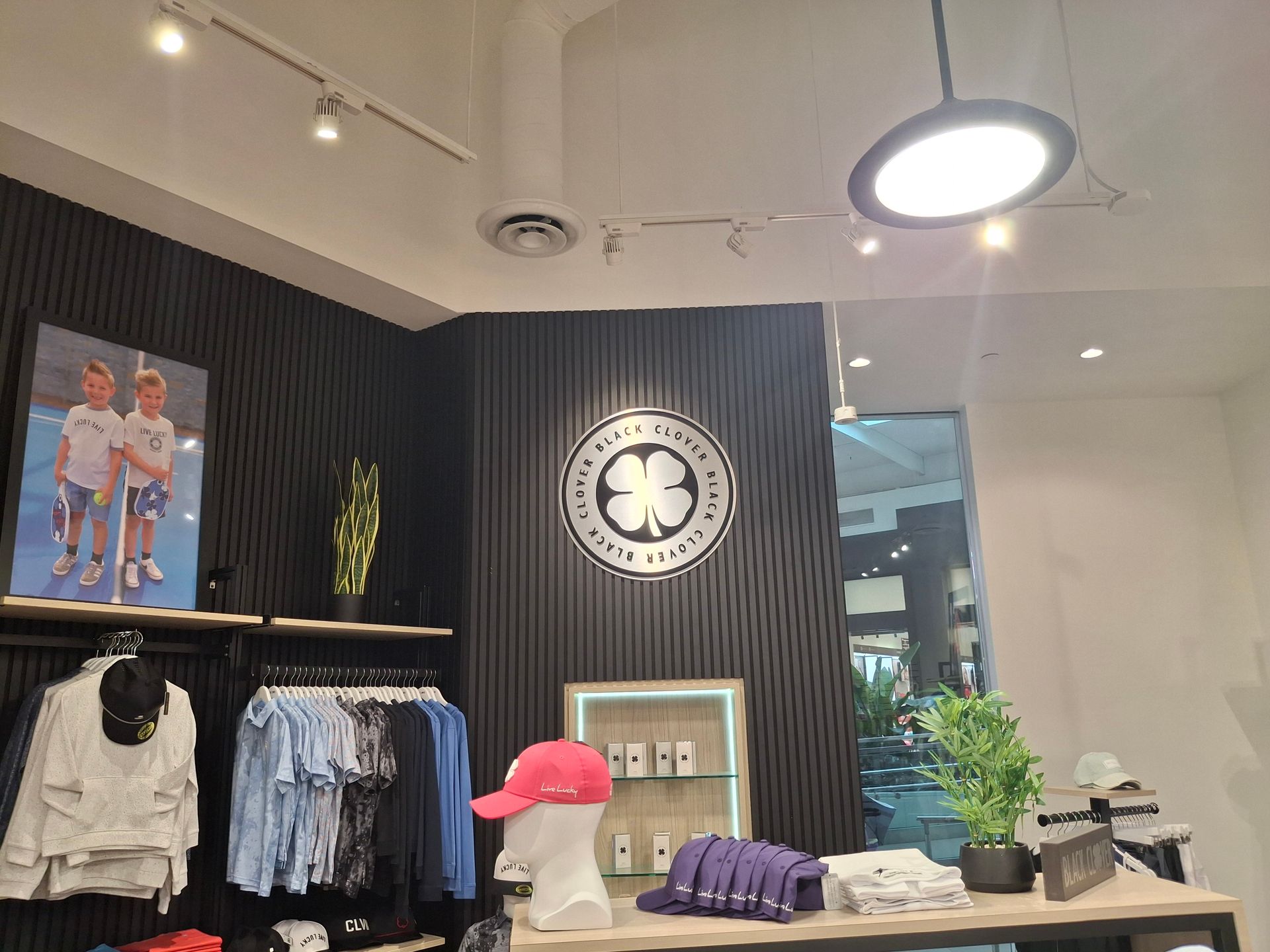 Clothing store interior with racks of shirts, a wall clock, display shelves, and a bright ceiling light
