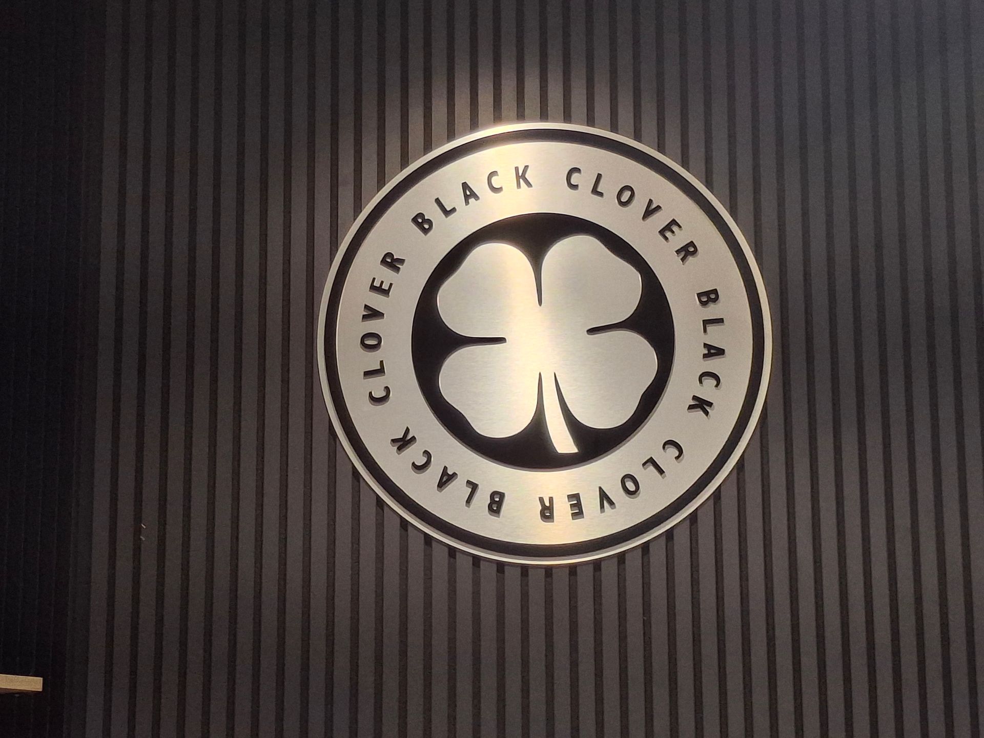Black Clover logo on a dark vertically striped wall, featuring a white clover emblem in a circular sign.
