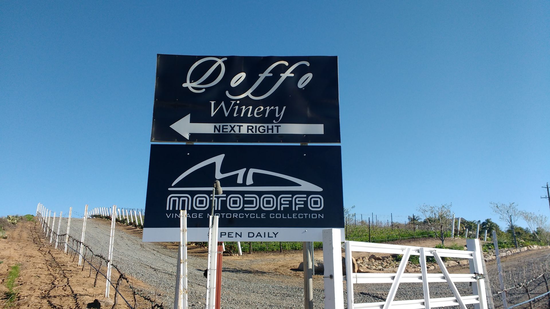 Roadside sign under blue sky pointing to a winery, with vineyards and a white fence nearby