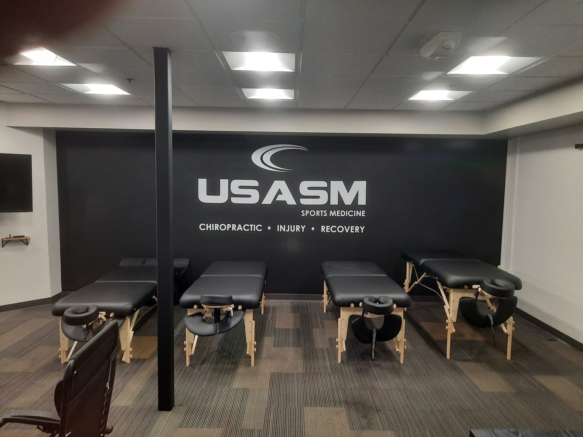 Massage therapy room with four treatment tables under ceiling lights and a USASM logo on the back wall