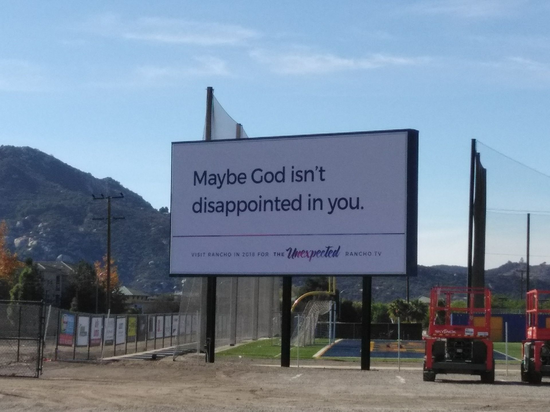 Billboard reading “Maybe God isn’t disappointed in you.” in a sunny outdoor field near mountains