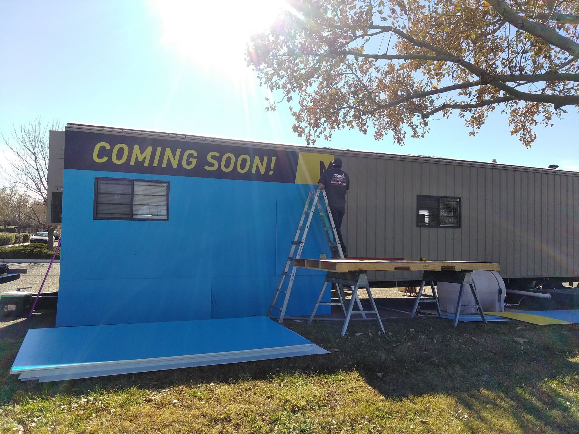 Blue “COMING SOON!” storefront with ladder and work table outside a trailer on a sunny day