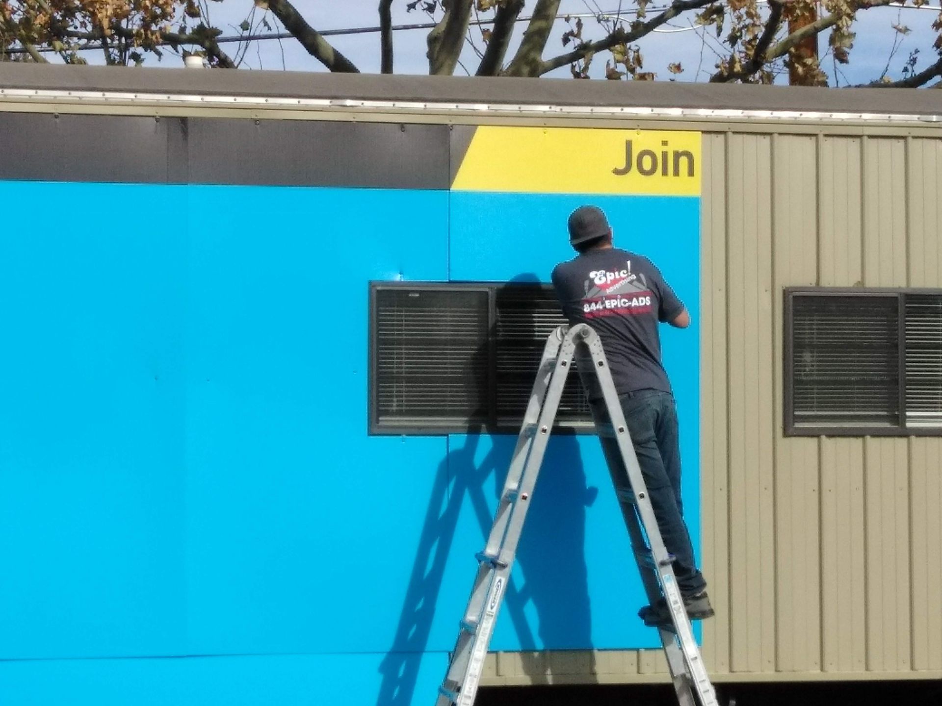 Worker on a ladder installing a sign on a bright blue wall next to a beige building.