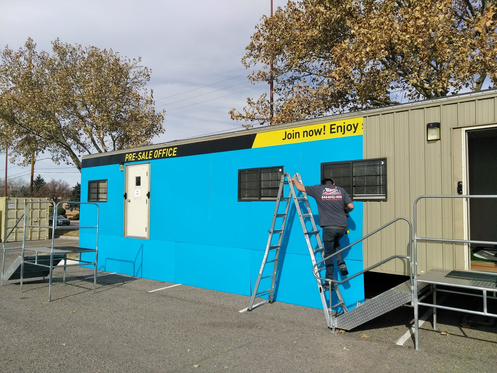 Worker on ladder painting a blue modular building with a yellow sign
