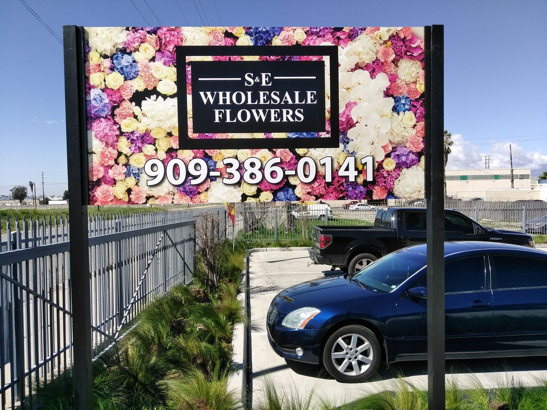 S.E. Wholesale Flowers sign with phone number, beside parked cars and a flower-filled fence.
