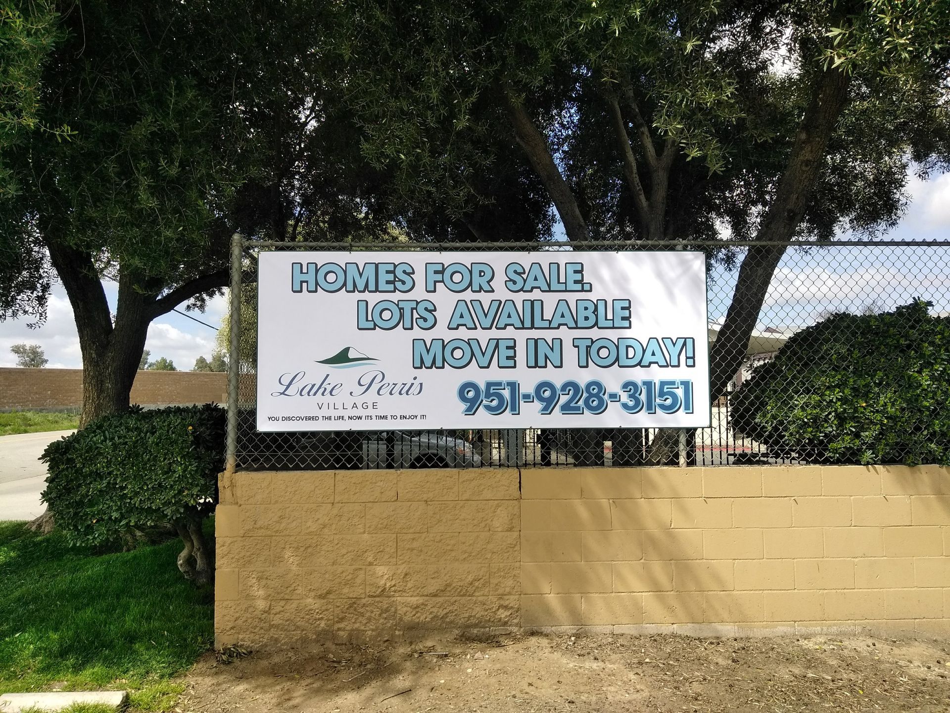 “Homes for Sale” sign on a tan wall under trees, with lot availability and phone number.