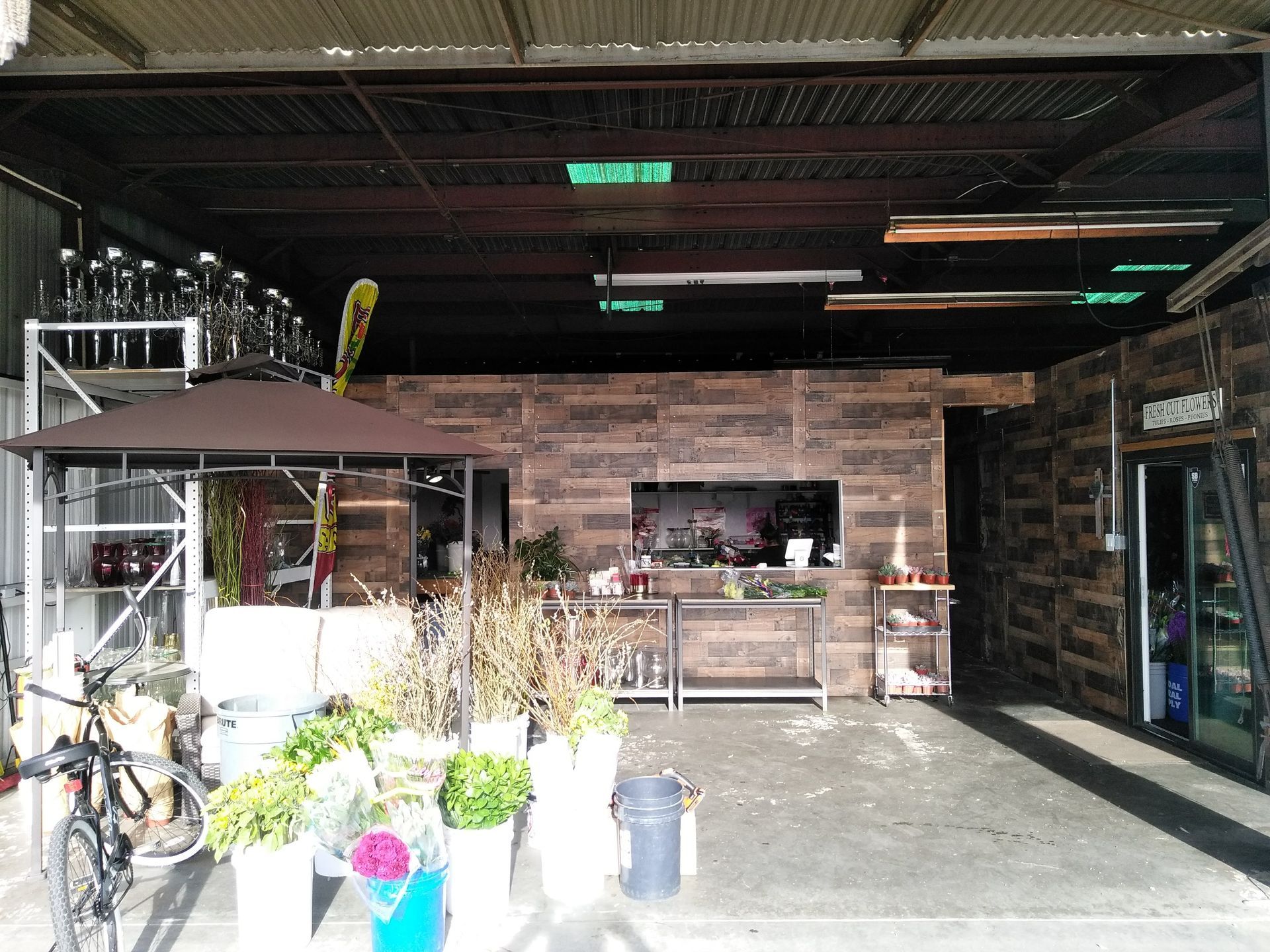 Open-air café storefront with potted plants, bicycles, and a wooden counter under a shaded roof