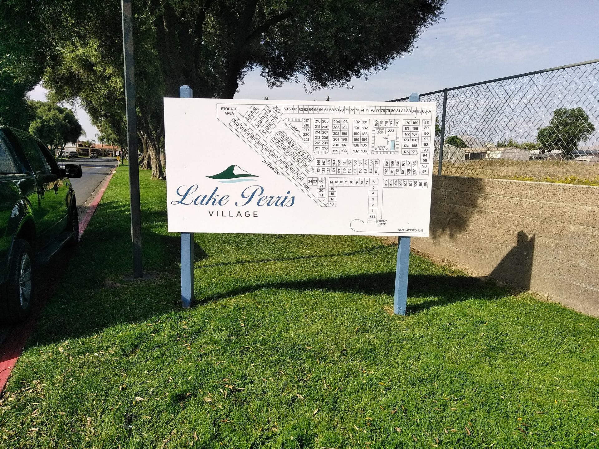 Lake Perris sign on blue posts beside a grassy path and chain-link fence under trees
