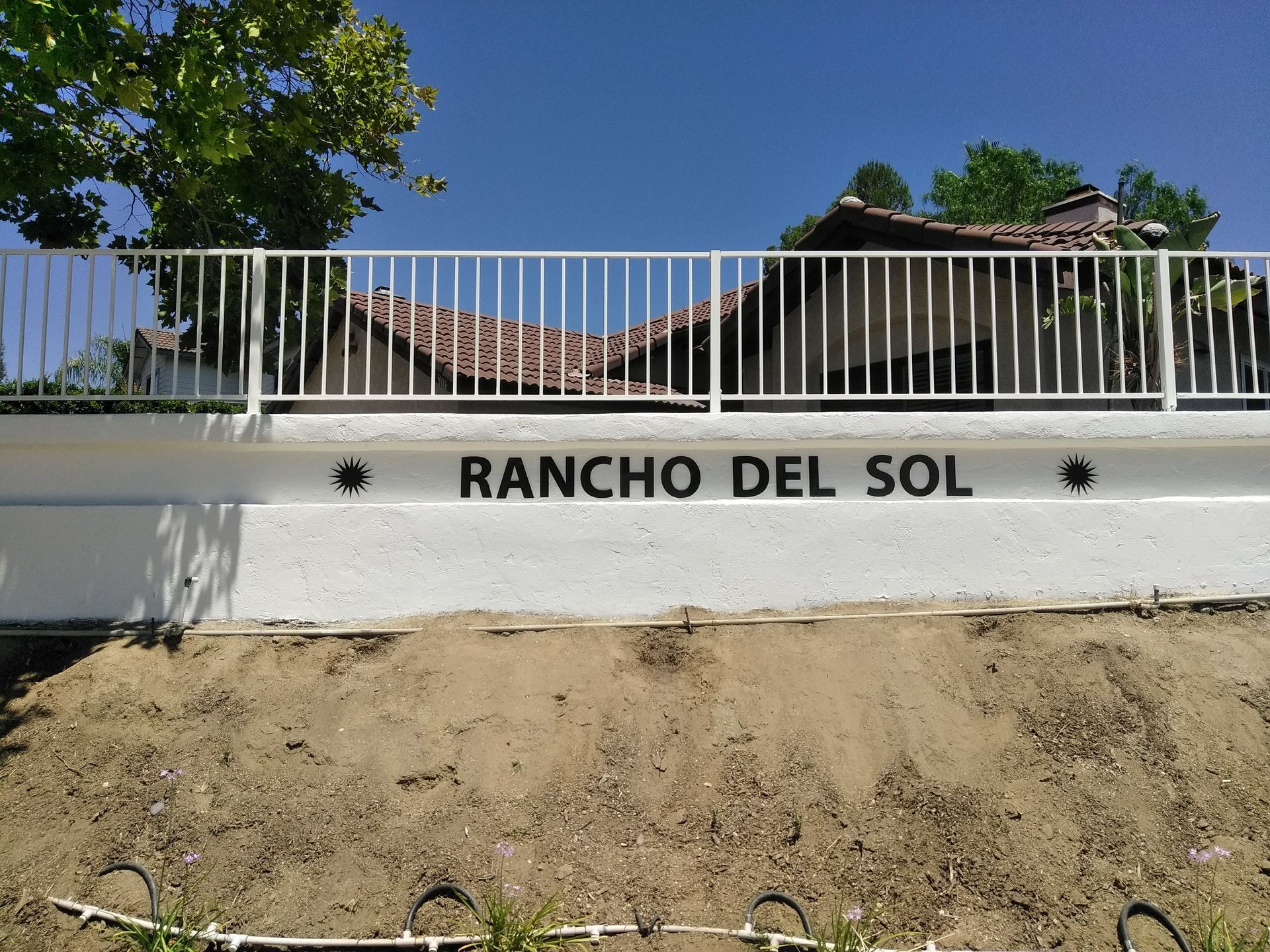 Rancho Del Sol sign on a white wall beneath a metal fence and trees under a blue sky