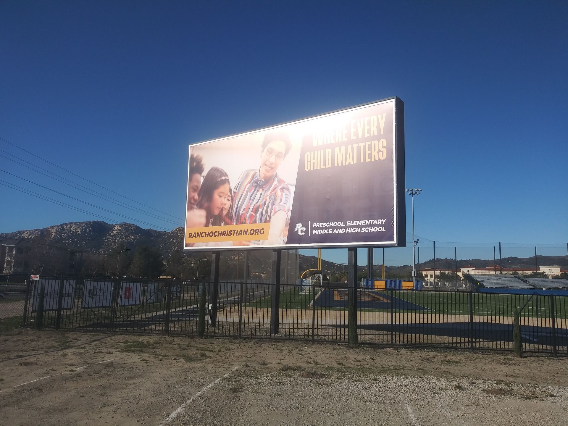 Large roadside billboard with bright movie-style posters beside a fenced field under a clear blue sky