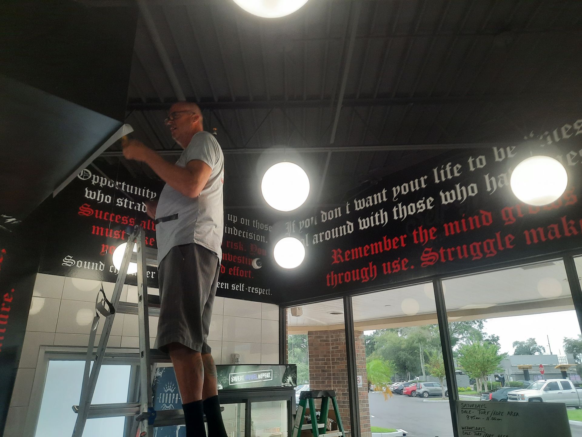 Man on ladder painting wall text inside a room with large windows and hanging lights.