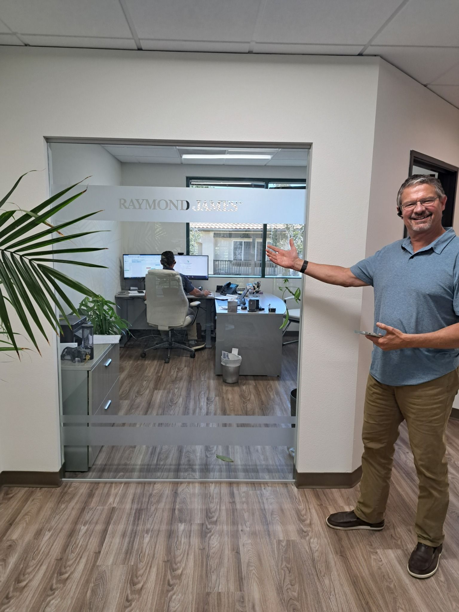 Man standing by glass office doorway, gesturing toward a conference room with desk and chairs inside.
