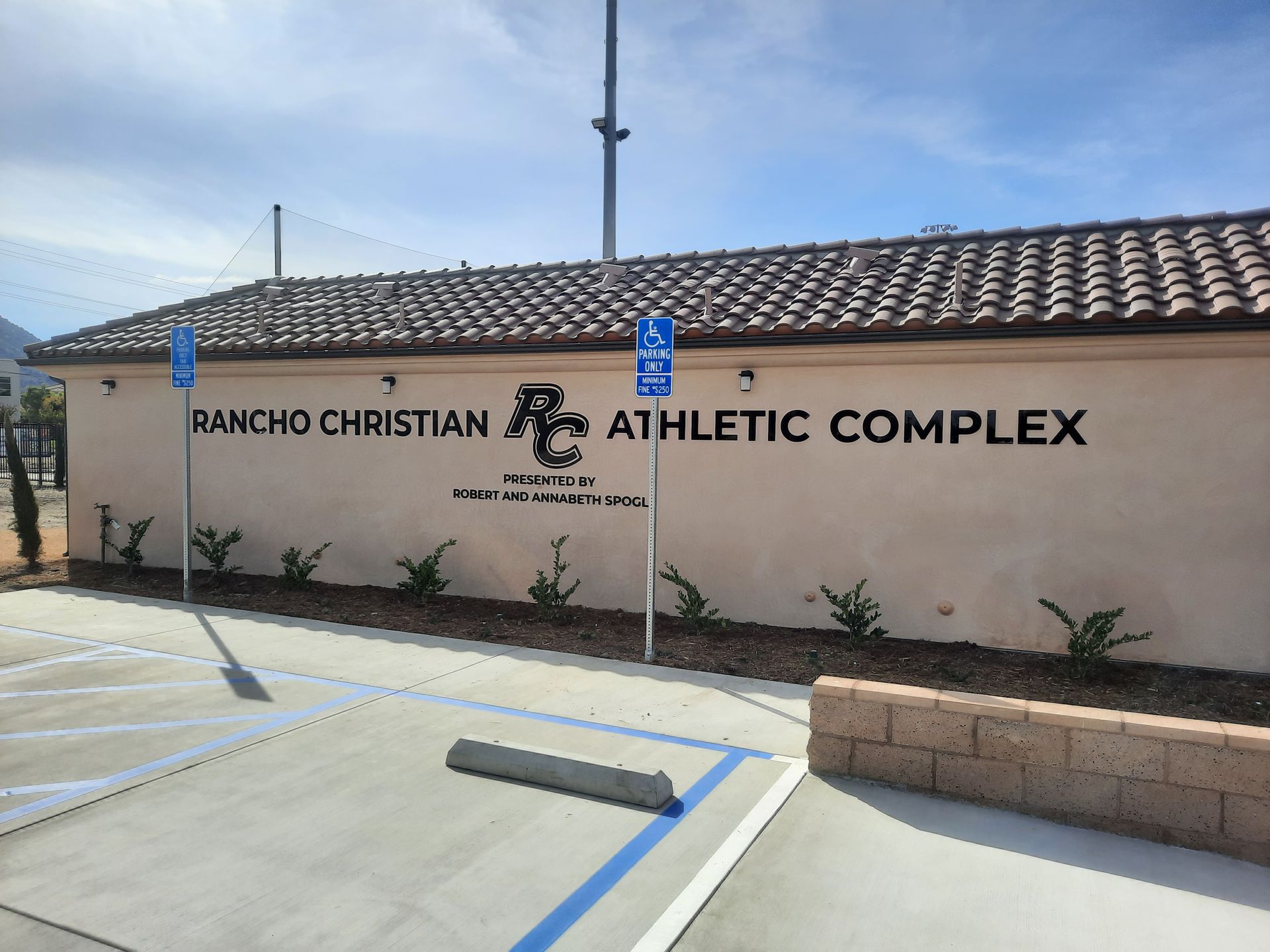 Ranch Christian Athletic Complex sign on a stucco wall with blue sky and parking lot in front