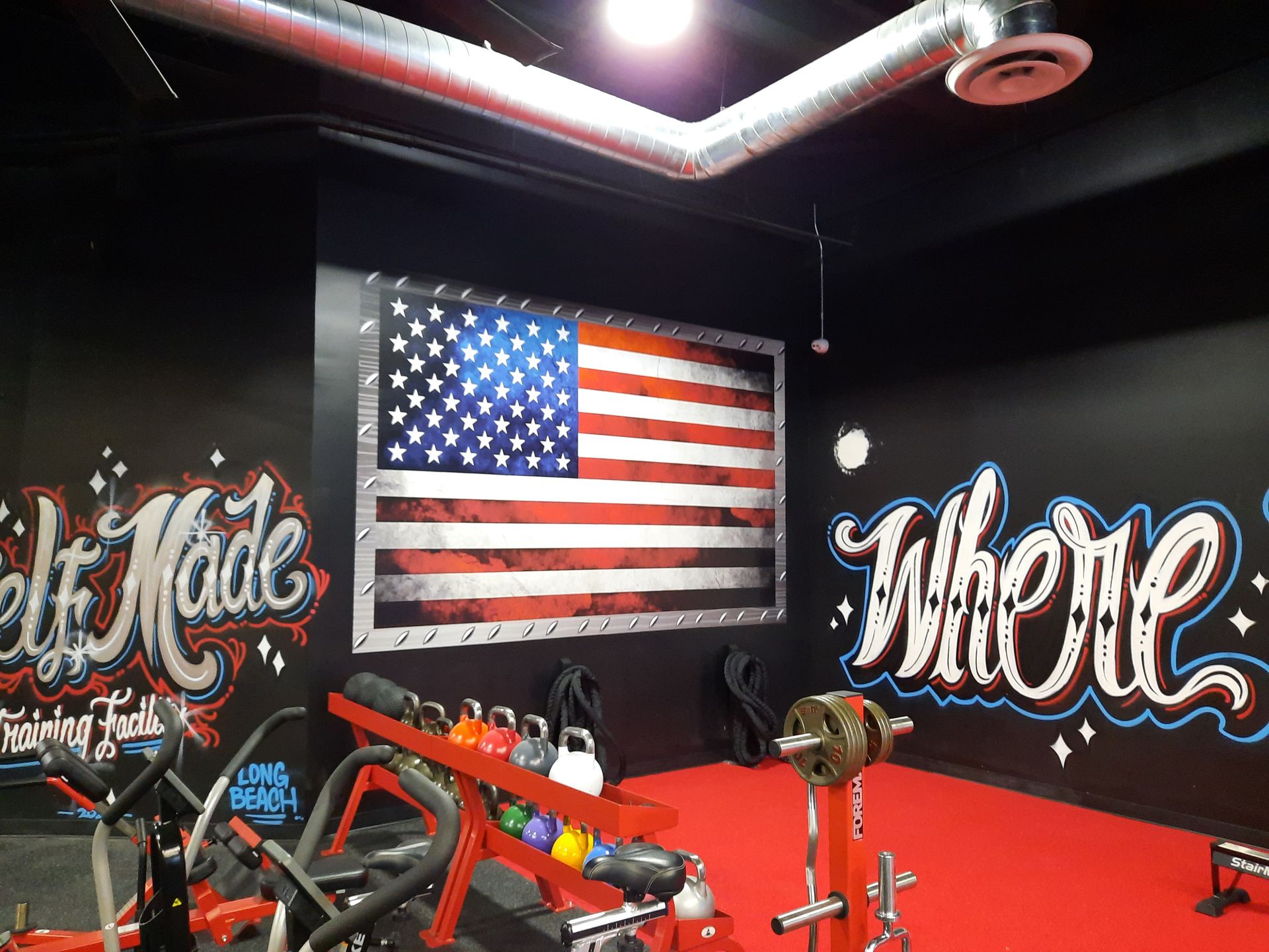 Gym room with large American flag mural, graffiti-style wall art, and red workout equipment on black walls.