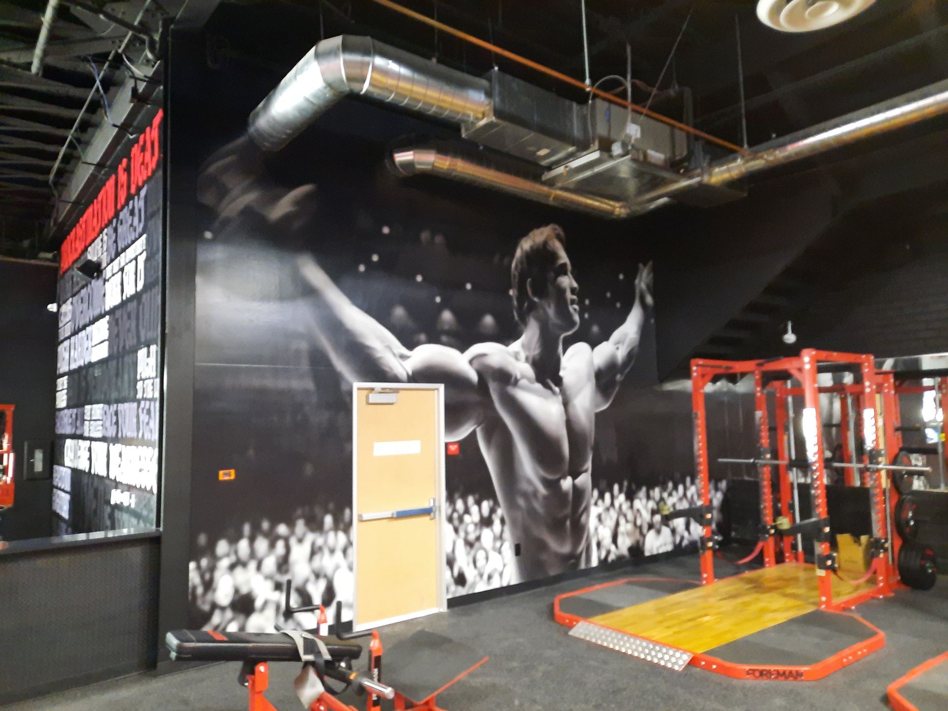 Gym interior with large black-and-white wall mural, red exercise equipment, and industrial ceiling ductwork.