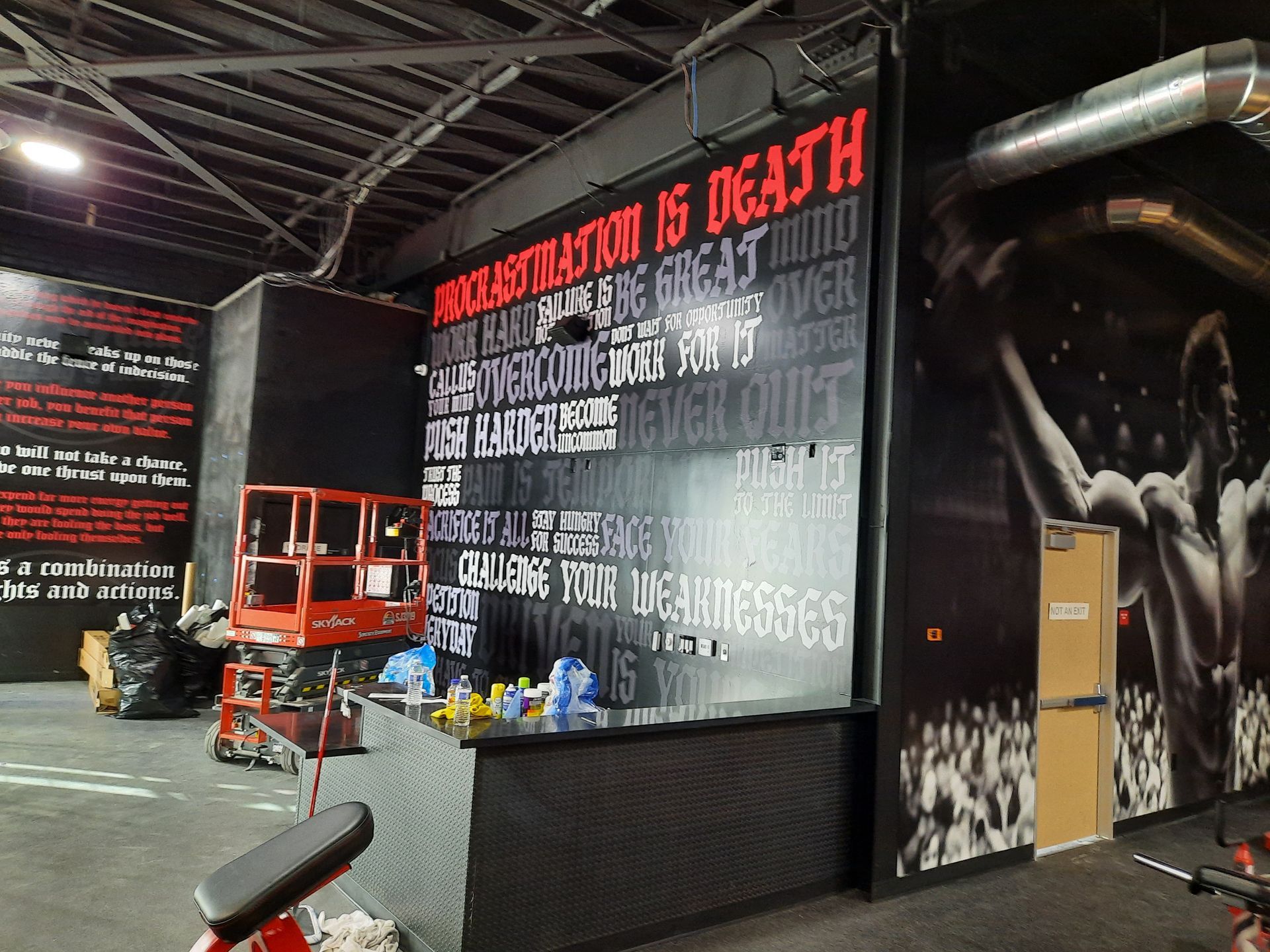 Indoor gym with black walls, workout equipment, and a bold red-and-white mural sign.