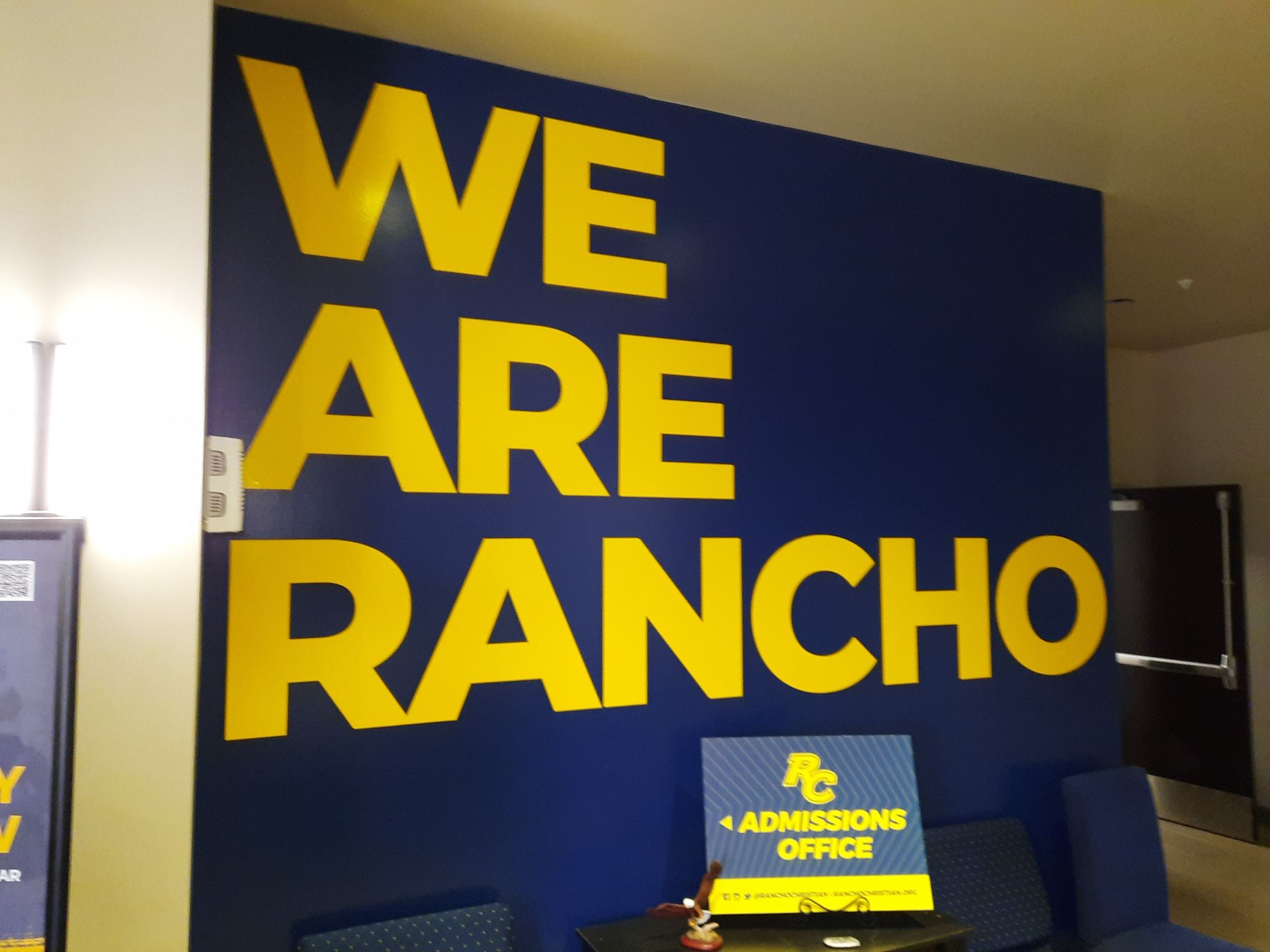 Large blue wall with bold yellow text reading “WE ARE RANCHO” in an indoor hallway