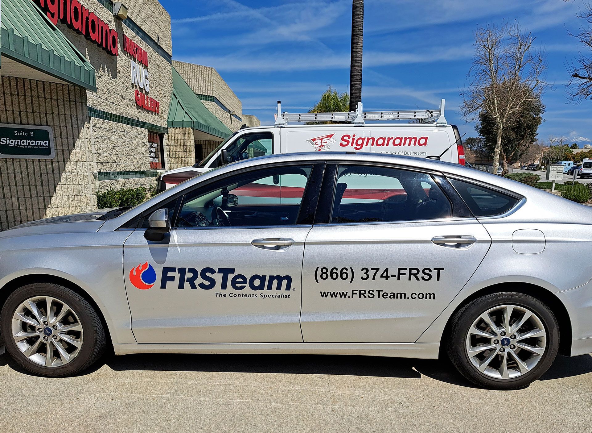 Silver FRSteam sedan parked outside a Signarama store with company logos on the side.