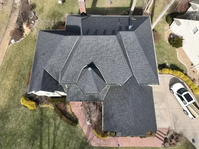 Overhead view of a house with a dark gray shingled roof, a brick walkway, and a white car in the driveway.
