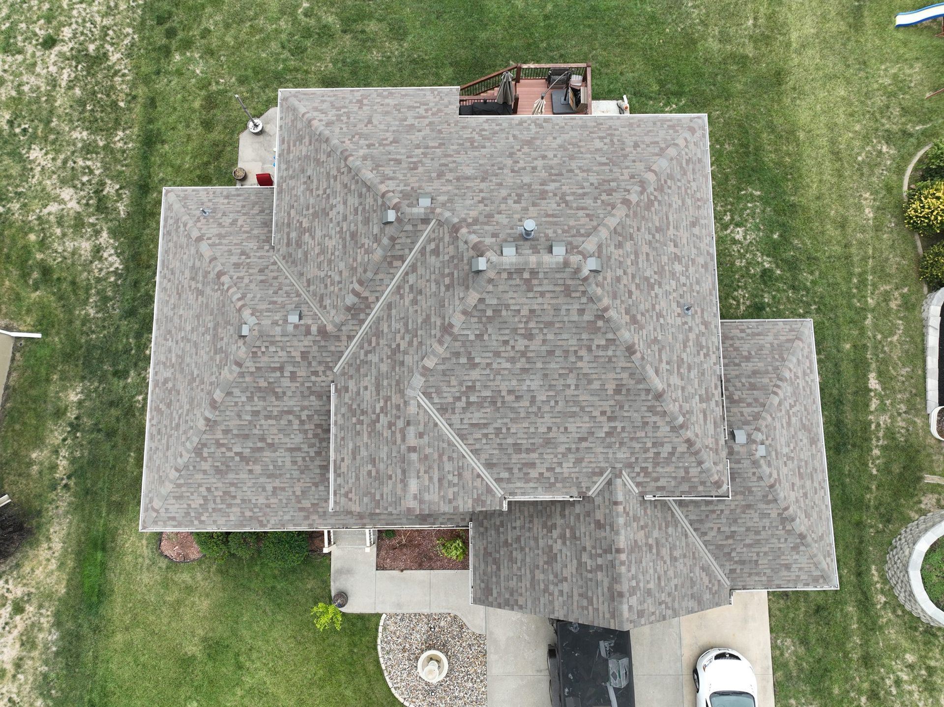 Overhead view of a house with a gray roof, surrounded by green grass. A driveway and car are visible.