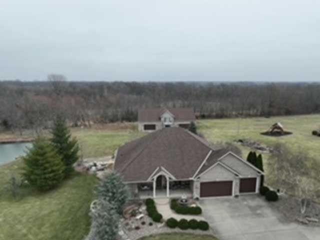House with brown roof and two-car garage on a grassy lot, with a pond and trees in the background.