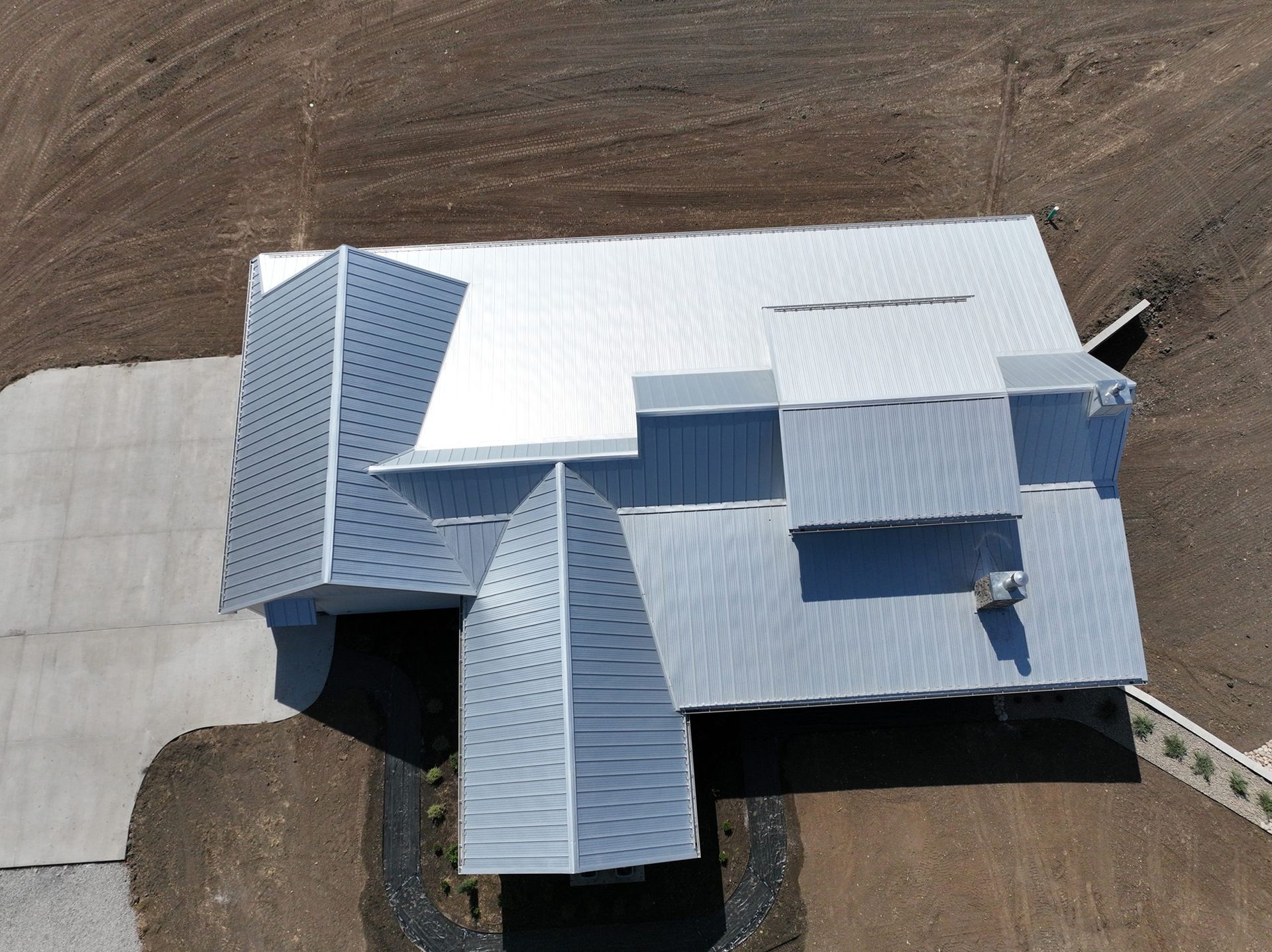 Aerial view of a modern house with a corrugated metal roof on a plot of land with gravel and a concrete driveway.