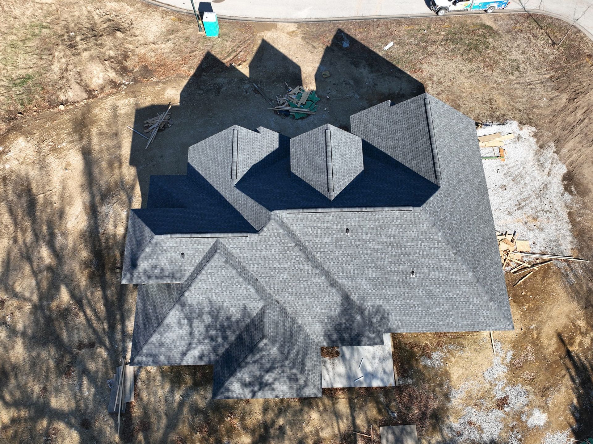 Aerial view of a dark gray shingled roof under construction, casting long shadows on the brown ground.