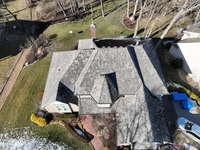Overhead view of a house with a gray shingle roof, chimney, and surrounding yard with some trees and patches of snow.
