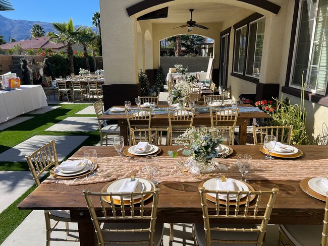 Elegant outdoor dining setup with long wooden tables, gold chairs, and floral centerpieces under a covered patio.