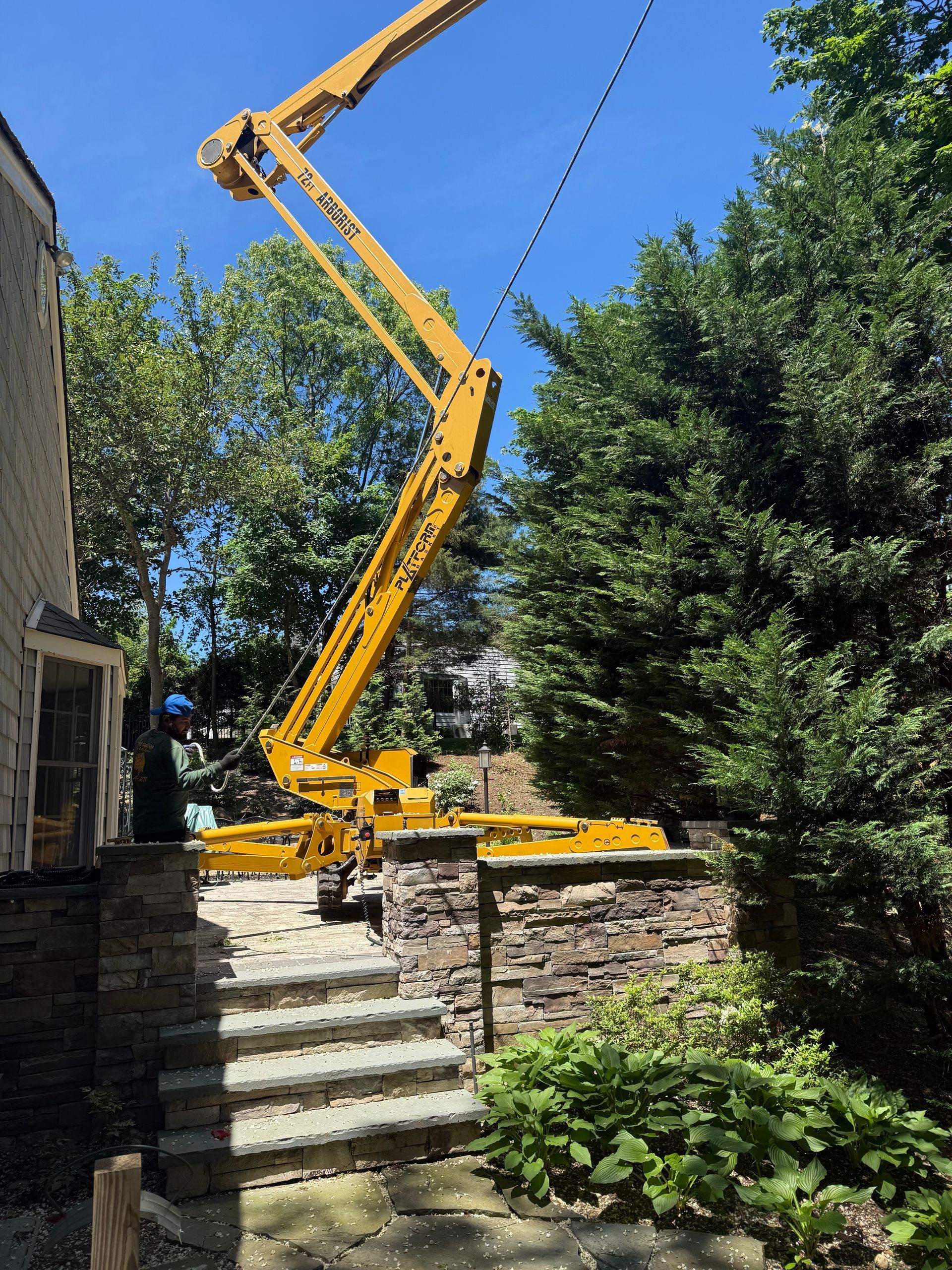 Yellow lift truck reaching toward power lines near a stone wall and house, under a blue sky.