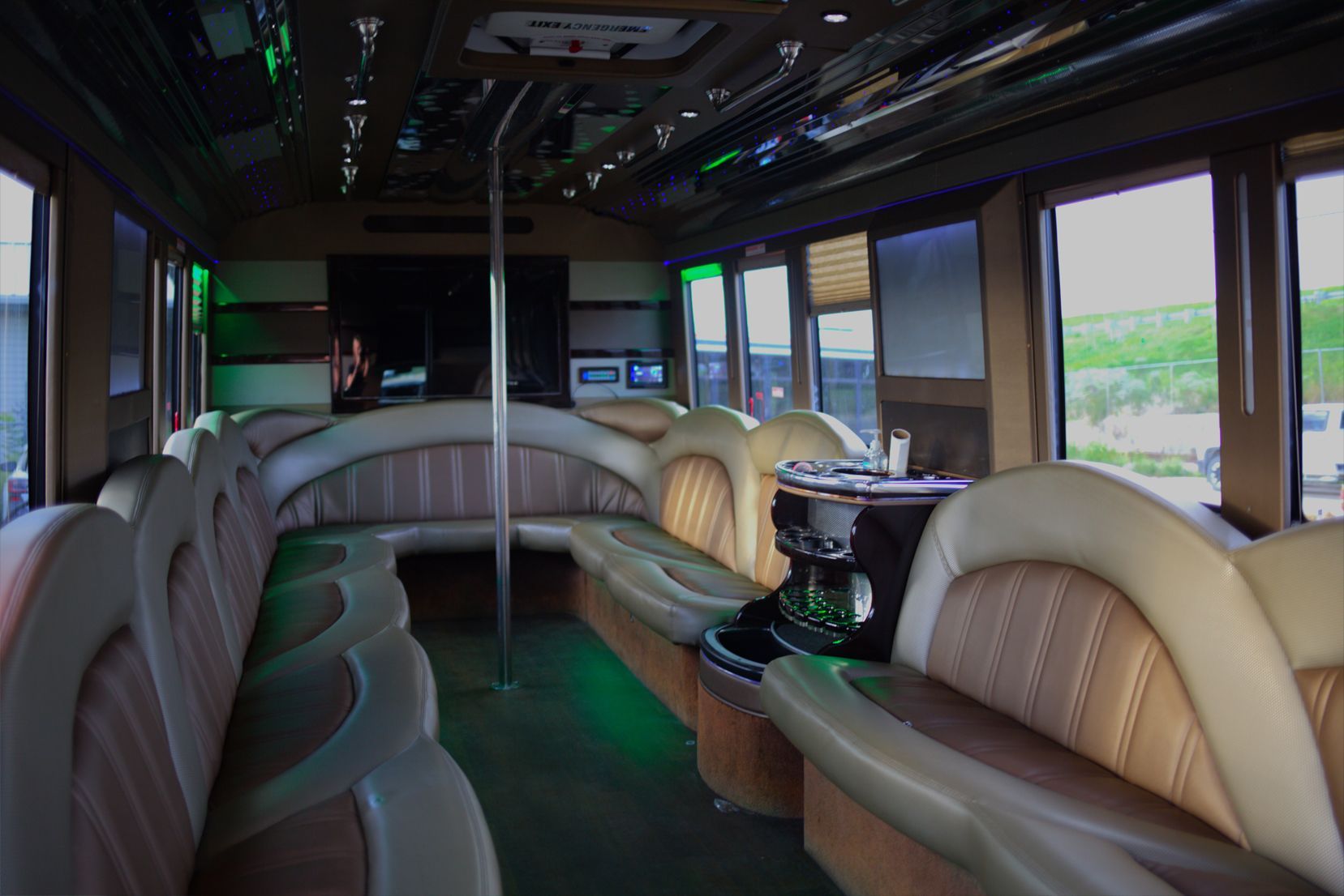 Interior of a party bus with cream and brown seating, a dance pole, and green accent lighting.