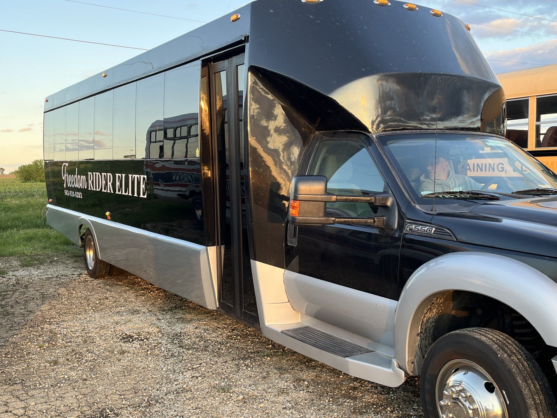 Black and silver luxury bus parked on gravel, with its door open. The bus is in a field under a cloudy sky.