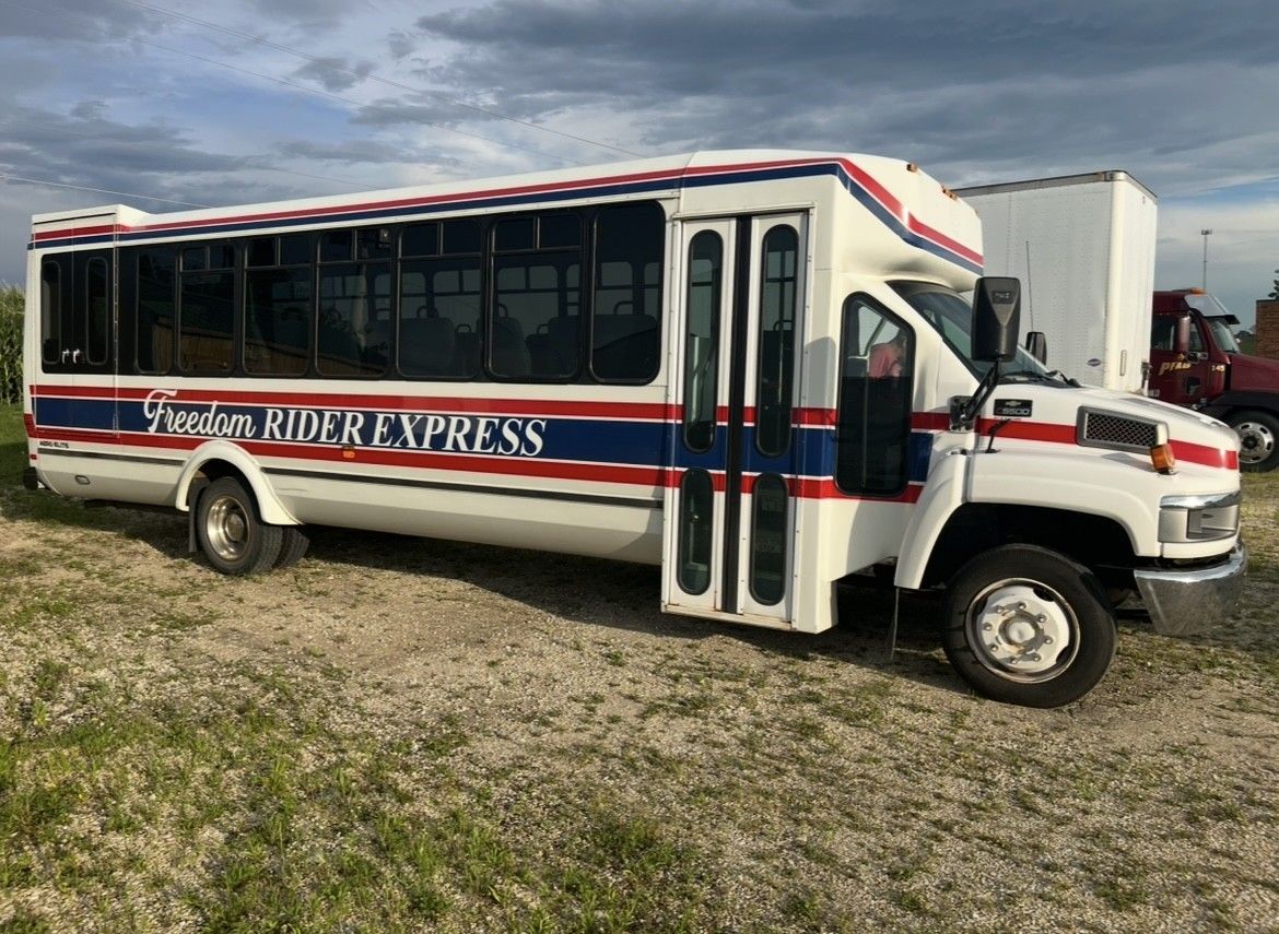 White passenger bus with red and blue stripes, parked on gravel. The bus reads