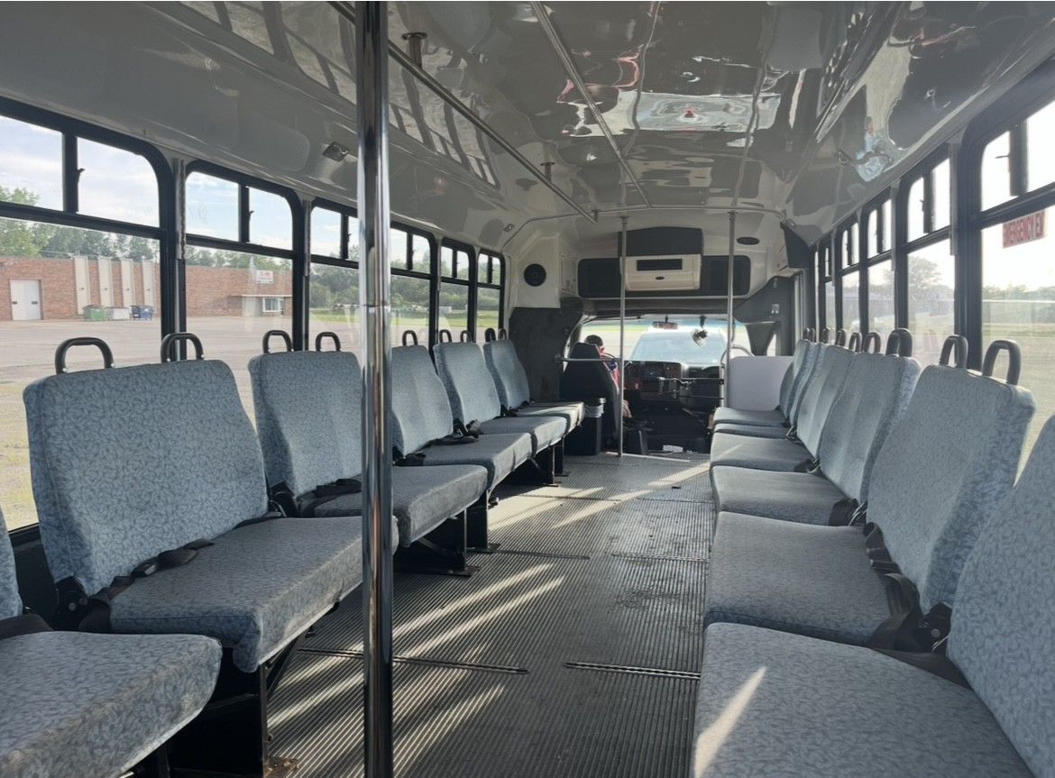 Interior of a bus with rows of blue seats, metal poles, and windows along the sides. A driver sits in the front.