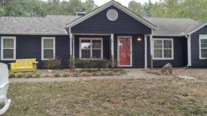 Single-story dark house with a red front door and small porch, framed by a grassy yard and trees.