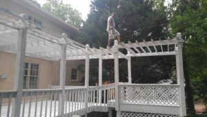 White pergola and lattice deck in a backyard, with a statue on top and trees in the background