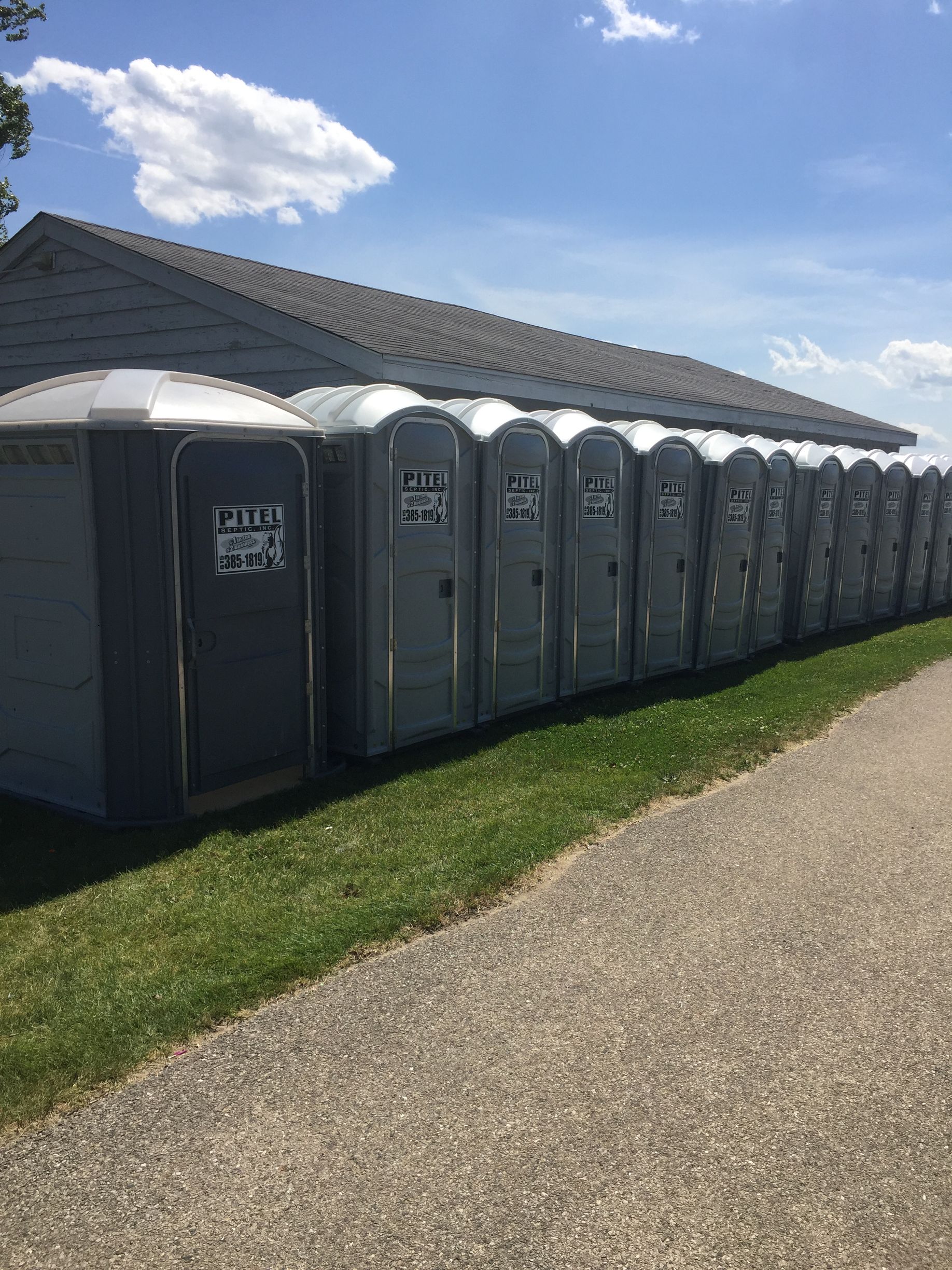 A row of grey portable toilets lined up on a grassy patch next to a building under a sunny, blue sky.