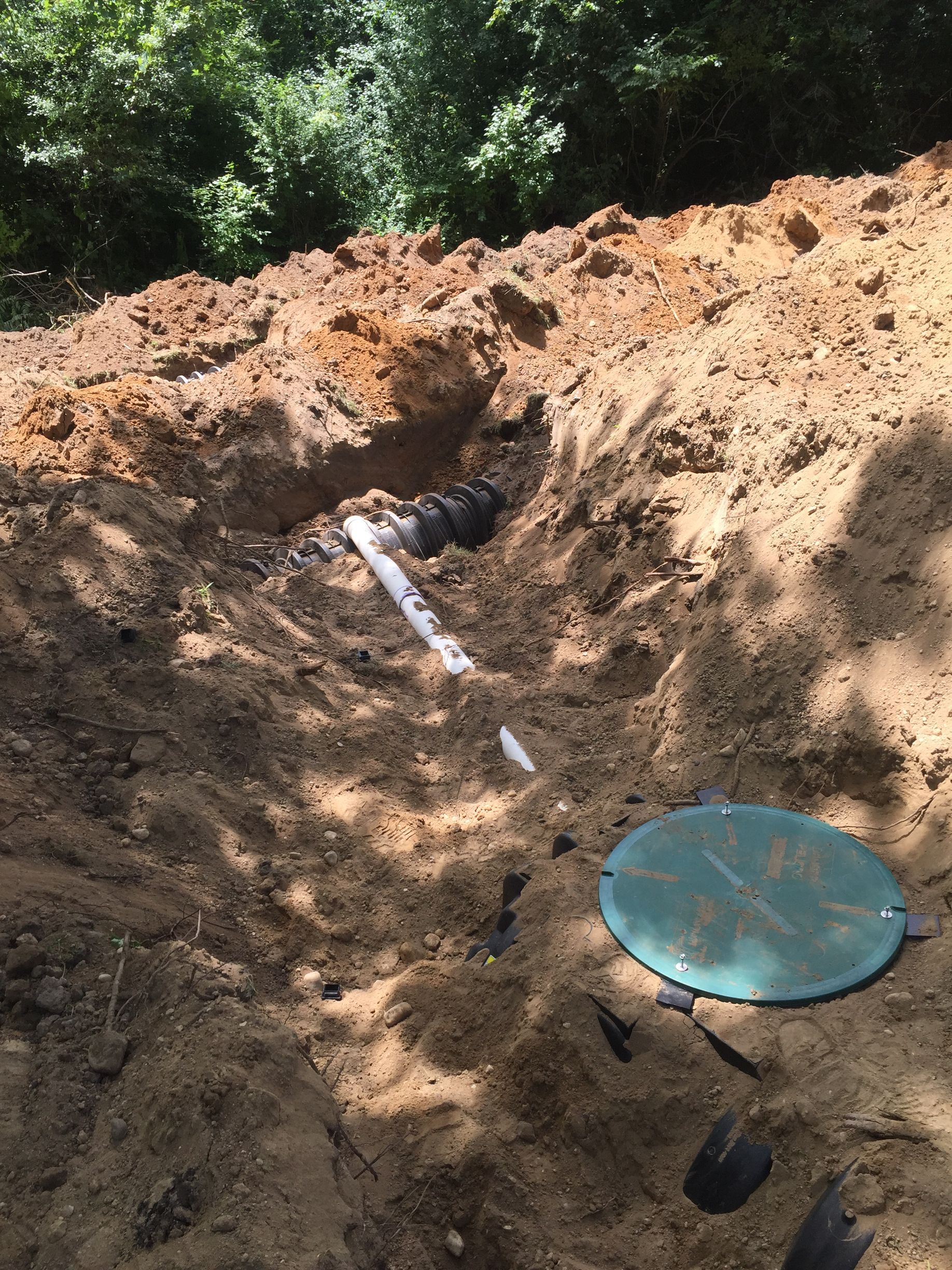A partially buried septic system tank with a green circular access lid and connecting pipes in a dirt trench.