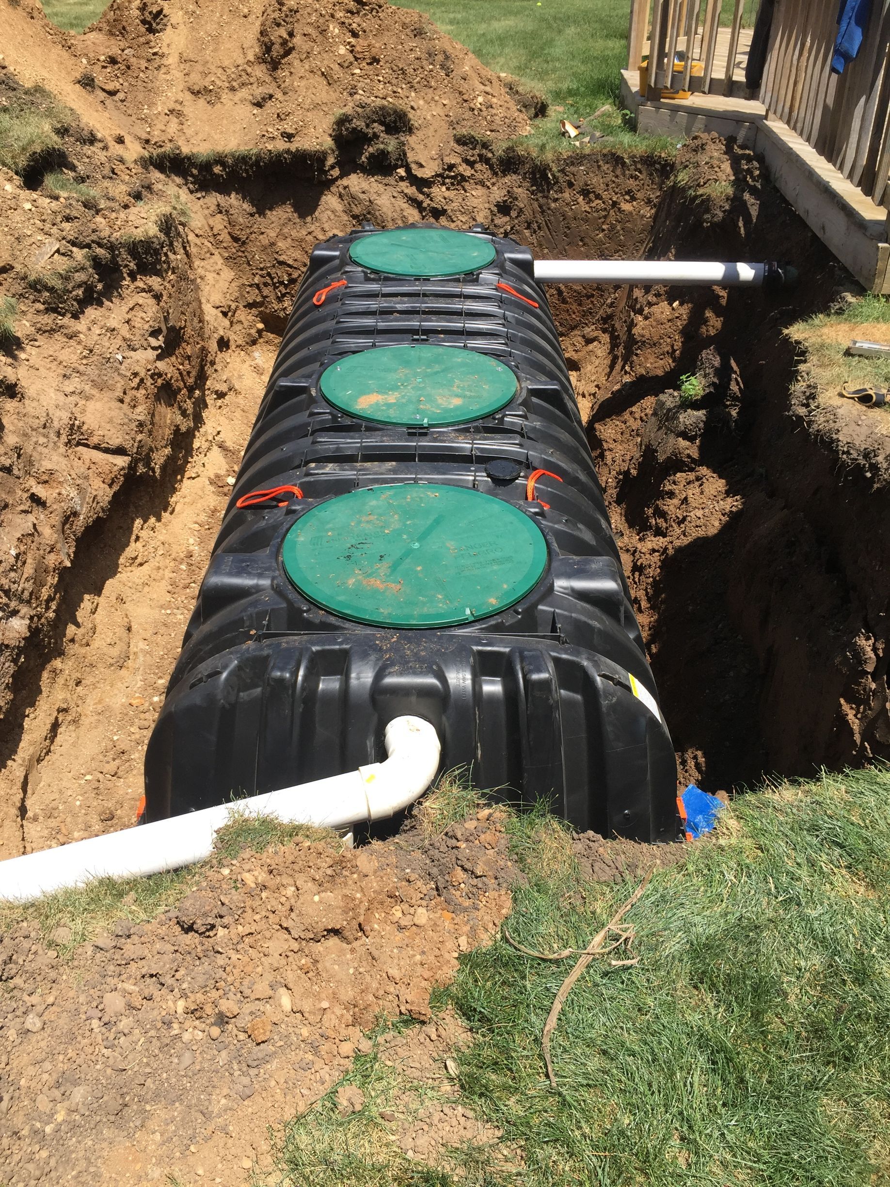 A newly installed black plastic septic tank with three green circular lids sits in a trench, connected to white pipes.