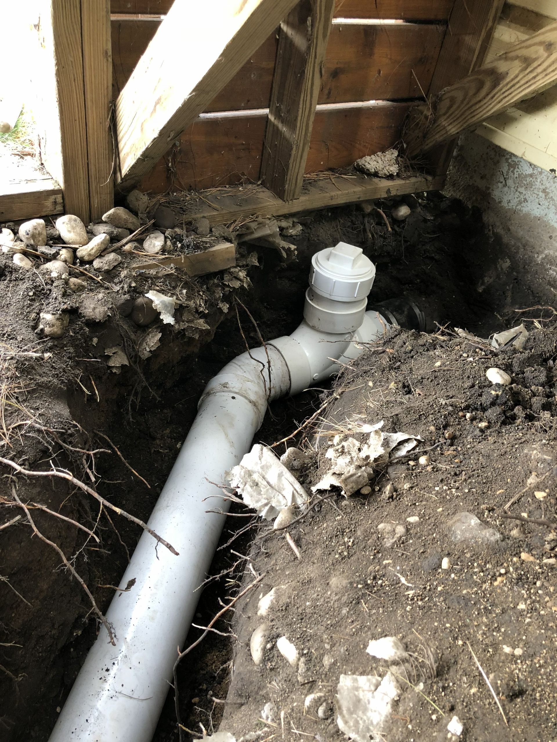 A gray PVC sewer cleanout pipe installed in a dirt trench beneath wooden porch supports.