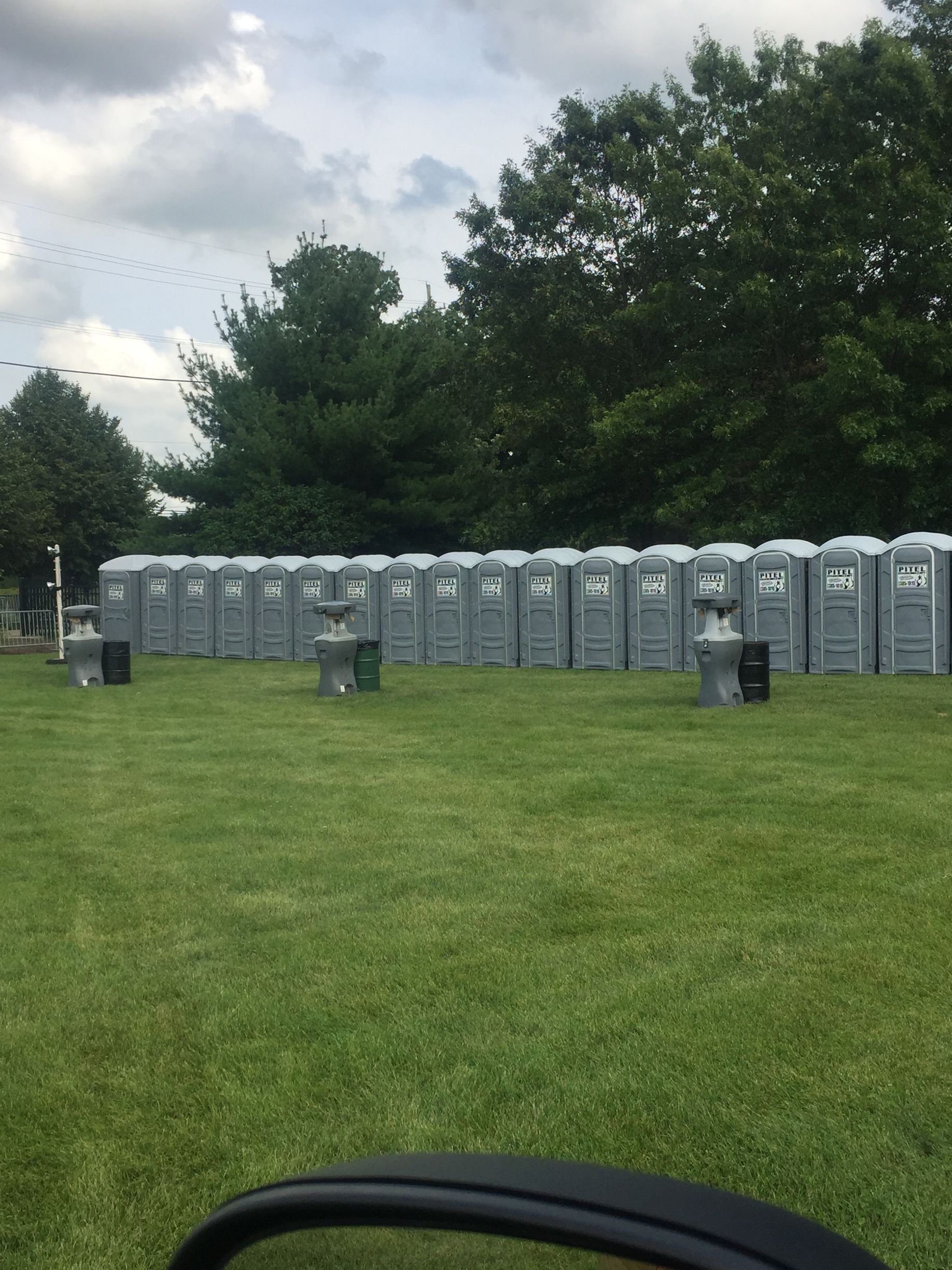A long row of gray portable toilets arranged in a line on a grassy field, with a few hand-washing stations in front.