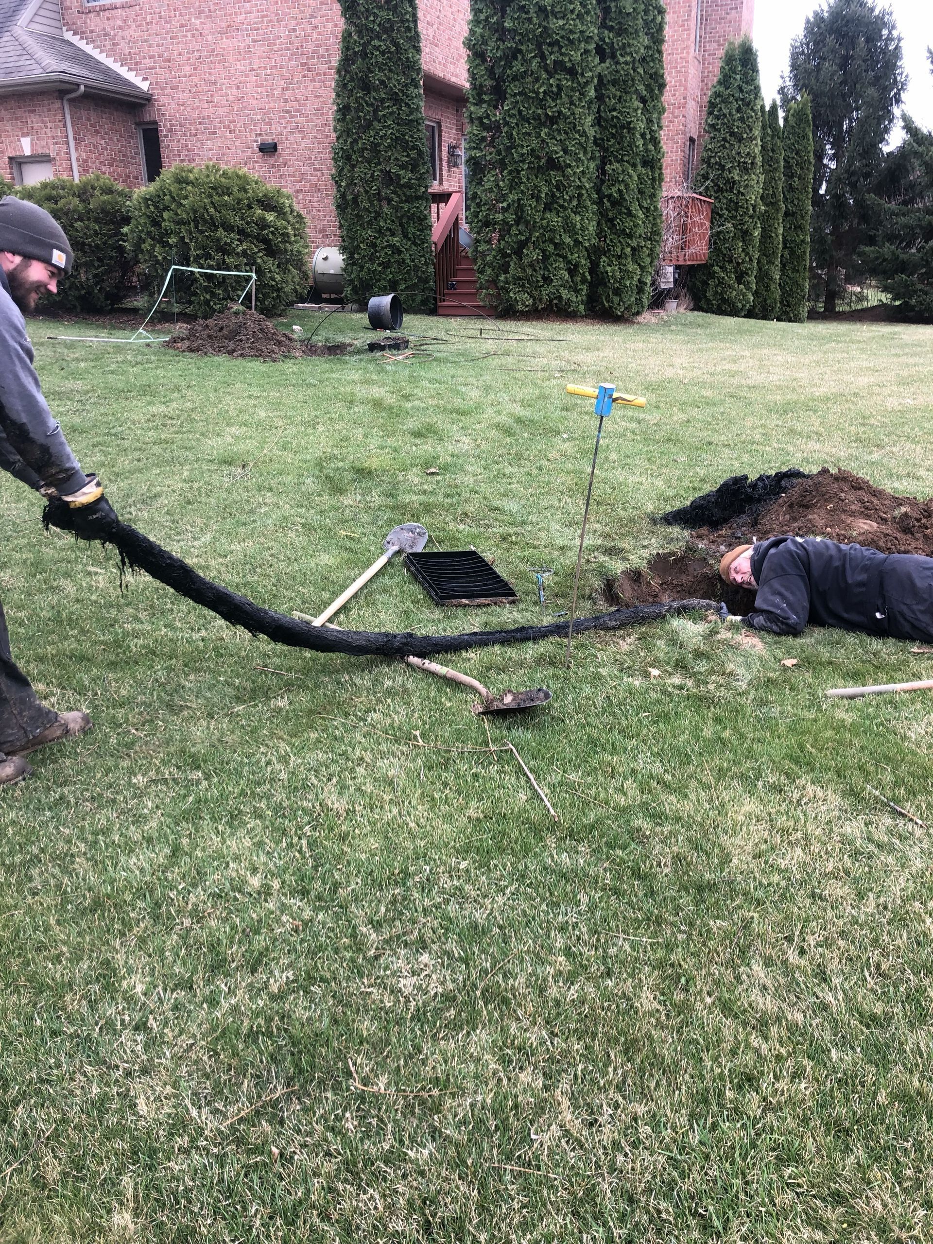 Two people work in a grassy yard, one pulling a long black drainage pipe toward the other who is lying in an open trench.